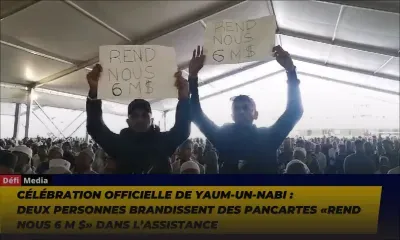 Célébration officielle de Yaum-Un-Nabi : deux personnes brandissent des pancartes «Rend Nous 6 M $» dans l’assistance
