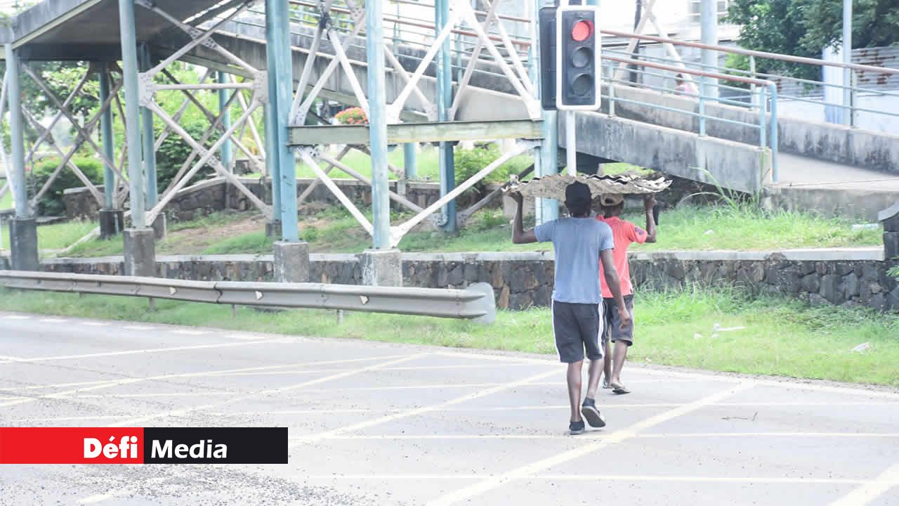 À Vallijee, deux hommes traversent la route avec des feuilles de tôle sur la tête qui les empêche de voir le trafic.